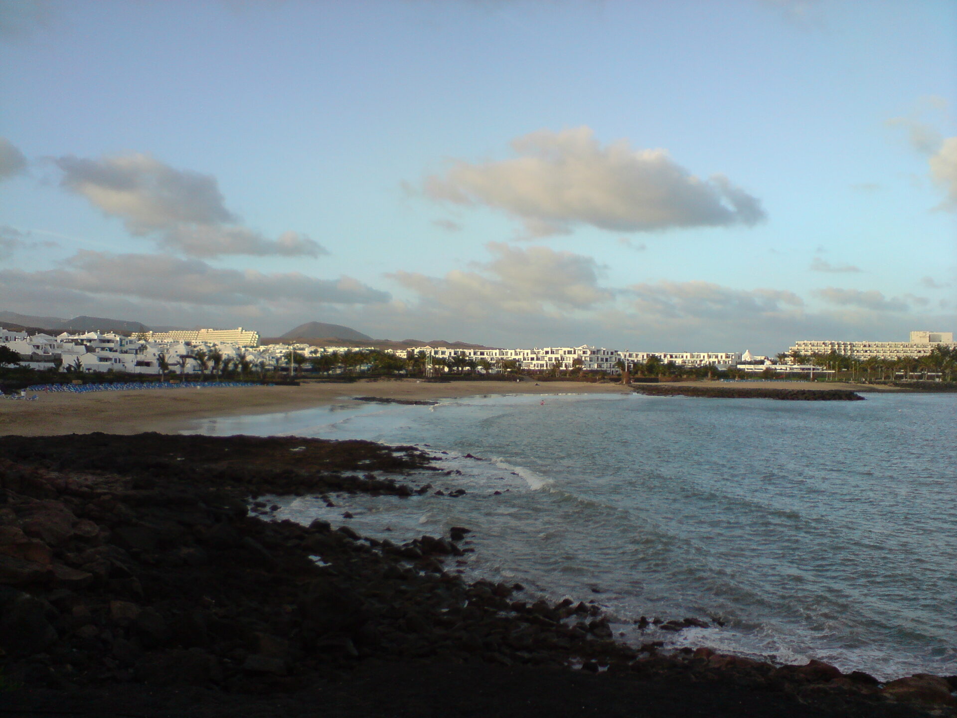 Bahía de Playa Las Cucharas en Costa Teguise, Lanzarote, con rocas volcánicas en primer plano al atardecer