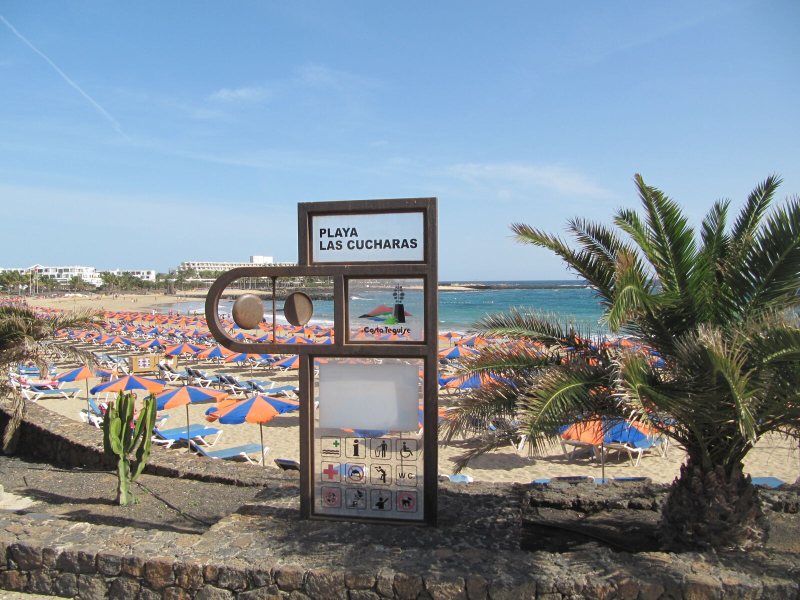 Cartel de Playa Las Cucharas en Costa Teguise, Lanzarote, con sombrillas naranjas al fondo