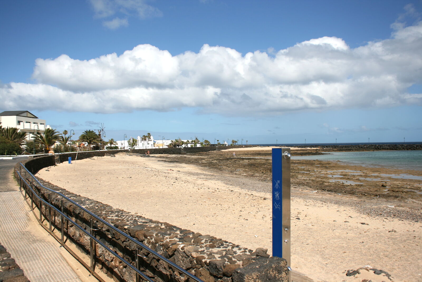 Playa de Los Charcos en Costa Teguise, Lanzarote, playa de arena con arrecife de roca volcánica descubierto en marea baja y rompeolas al fondo